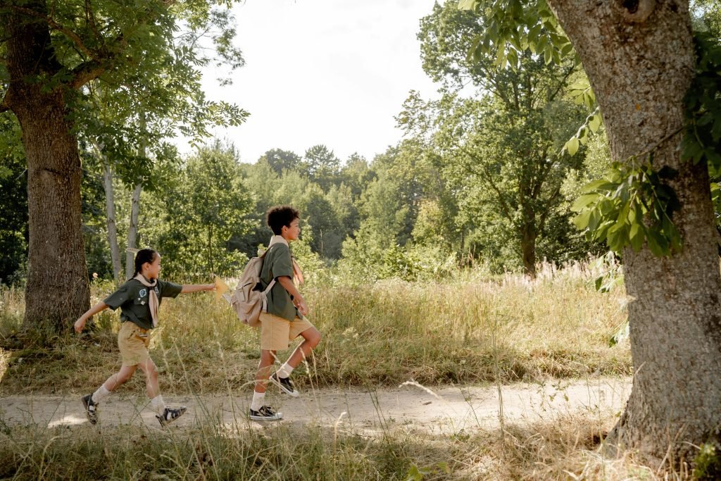 Two children in scout uniforms hiking along a forest path on a sunny day.